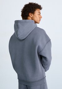 Sweatshirt à capuche gris en tissu doux, coupe oversized et détails de couture visibles. Comprend une poche kangourou et des poignets côtelés.