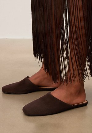 Picioare purtând pantofi maro închis, fără șiret, cu vârf ascuțit, lângă o țesătură lungă cu franjuri întunecați ce atârnă aproape de glezne.