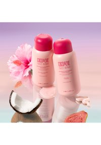 Två rosa schampo- och balsamburkar, märkta "Coco & Eve Sweet Repair," bredvid en kokosnötshalva och en guavaskiva på en reflekterande yta.