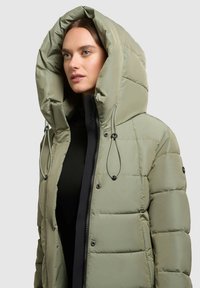 Pufota jaka gaisā olīvu zaļā krāsā ar lielu kapuci, ar melnu rāvējslēdzēju, šņorēm un apaļām pogām uz piedurknēm.