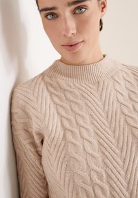 Beige Strickpullover mit hohem, geripptem Kragen, der ein Kabelmuster und eine strukturierte Oberfläche aufweist. Der Stoff wirkt weich und gemütlich.