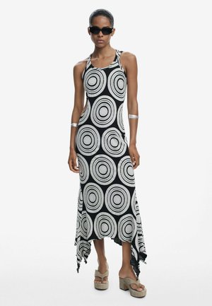 Schwarz-weißes ärmelloses Kleid mit kreisförmigen Mustern, asymmetrischem Saum, aus einem leichten Stoff gefertigt. Model trägt Wedge-Sandalen.