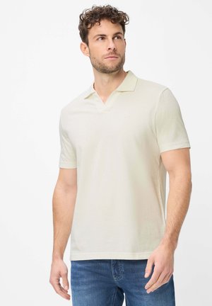 Man met kort krullend haar, gekleed in een crème kortemouwe polo en blauwe spijkerbroek, kijkt iets naar rechts.