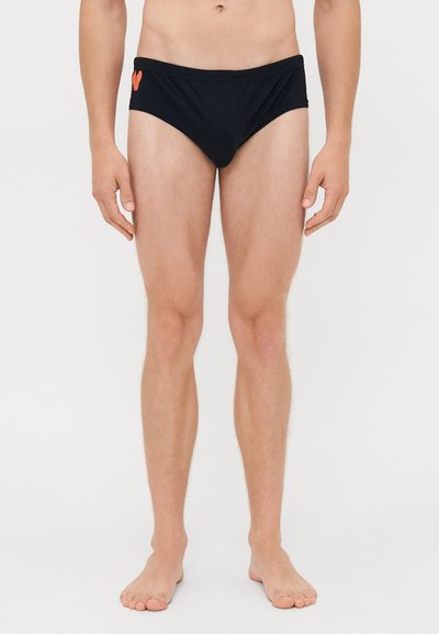 Male model standing barefoot wearing black swim briefs with an orange logo on the left side against a plain white background.