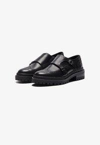 Black leather shoes with two straps, decorative brogue detailing, and a chunky rubber sole. Smooth texture with contrasting stitching accents.