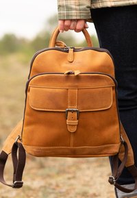 Mochila de cuero marrón con forma rectangular, asa en la parte superior, dos correas ajustables, bolsillo frontal y detalle de hebilla metálica.