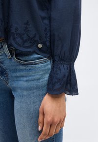 Μπλούζα σε ναυτικό μπλε με μακριά μανίκια, κεντημένα λουλουδένια σχέδια και κυματοειδείς άκρες, συνδυασμένη με μπλε τζιν τζιν, χέρι ακουμπισμένο δίπλα.