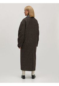 Brauner, strukturiertes Langmantel mit einem schwarzen Lederkragen, weiten Ärmeln und seitlichen Schlitzern, kombiniert mit hellen Socken und schwarzen Schuhen.
