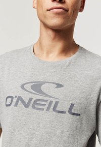 Šedé bavlněné tričko s tmavě šedým logem "O'Neill" a stylizovaným vlnovým designem. Výstřih je kulatý a látka vypadá měkce.