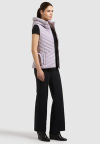Femme portant un gilet matelassé à capuche violet clair par-dessus une chemise noire, un pantalon large noir et des bottines compensées noires, debout de côté.