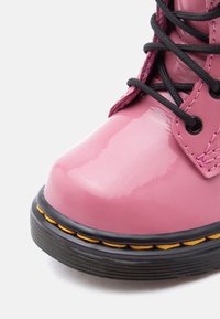 Γυαλιστερό ροζ δερμάτινο μποτάκι με μαύρη σόλα, κίτρινα ραφές και μαύρα κορδόνια δεμένα σε σταυρωτό μοτίβο σε λευκό φόντο.