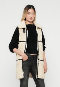 Ung kvinna klädd i svart fuzzy tröja, krämfärgad sherpa-väst med svarta detaljer och svarta texturerade byxor, stående med en hand i fickan.