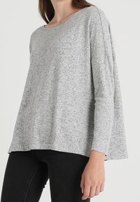 Graues Langarmshirt mit Rundhalsausschnitt und leicht lockerer Passform. Strukturierter Stoff mit meliertem Muster, kombiniert mit dunklen Jeans.