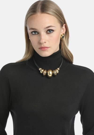 Gul statement halskæde med en blanding af glatte og teksturerede perler, på en sort læder-snøre, båret over en sort turtleneck sweater.