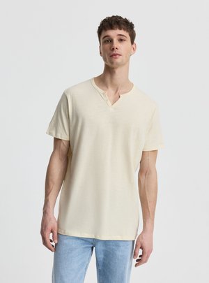 Młody mężczyzna w jasnobeżowej koszulce henley i niebieskich dżinsach stoi na jednolitym jasnym tle, zwrócony twarzą do przodu w zrelaksowanej pozycji.