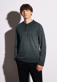 Langärmliges Henley-Shirt in Dunkelgrün, aus strukturiertem Stoff, mit einer Knopfleiste mit drei Knöpfen und einem kleinen aufgedruckten Logo auf der Brust.