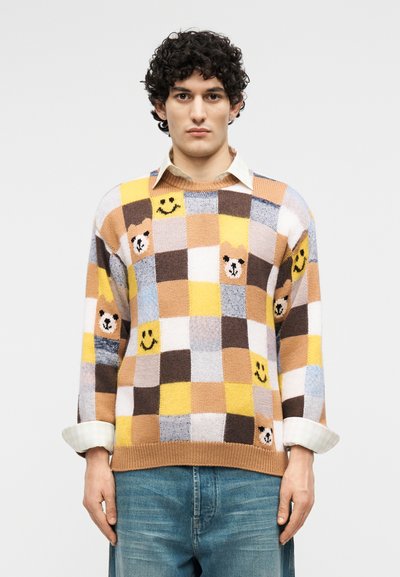Άνδρας που φοράει πολύχρωμο καρό πουλόβερ με πρόσωπα αρκούδων και μοτίβα χαμόγελου, σε συνδυασμό με λευκό πουκάμισο και μπλε τζιν.