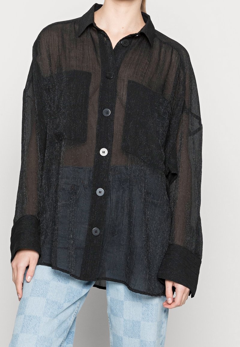 Femme portant une chemise noire ample et transparente à boutons avec deux poches avant, par-dessus un haut noir, associée à un jean à carreaux bleu clair.