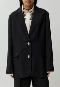 Zwarte blazer met een getailleerd ontwerp, voorzien van drie knopen, twee voorzakken en een zachte textuur. Draagt over een beige blouse met borduurwerk.