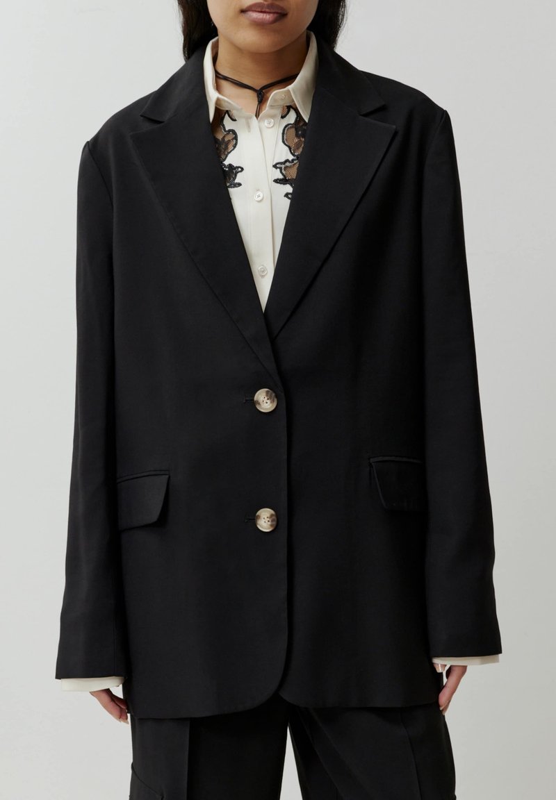 Zwarte blazer met een getailleerd ontwerp, voorzien van drie knopen, twee voorzakken en een zachte textuur. Draagt over een beige blouse met borduurwerk.