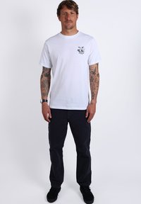 T-shirt en coton blanc avec un petit graphique noir sur la poitrine gauche, associée à un pantalon sombre et des chaussures noires à enfiler. Le modèle se tient droit.