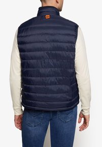 Marineblå oppustelig vest med høj krave og horisontalt quiltet mønster; har et orange logo-mærke på ryggen. Båret over en lys skjorte.