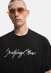 T-shirt nera con scollatura rotonda, caratterizzata da testo bianco scritto a mano. Il modello indossa occhiali da sole con tonalità arancione e montatura in metallo sottile.