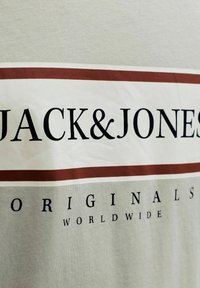 Μπεζ ύφασμα με τη λέξη "JACK & JONES" σε μεγάλα μαύρα γράμματα, περιτριγυρισμένο από δύο οριζόντιες καφέ ρίγες και με την ένδειξη "ORIGINALS WORLDWIDE" από κάτω.