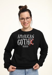 Donna sorridente con occhiali in un felpa nera con la scritta "American Goth X Wednesday," abbinata a jeans azzurri su uno sfondo bianco.