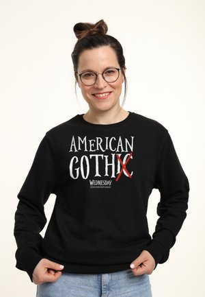 Usmívající se žena s brýlemi v černé mikině s nápisem "American Goth X Wednesday", které má na sobě světlé modré džíny na bílém pozadí.