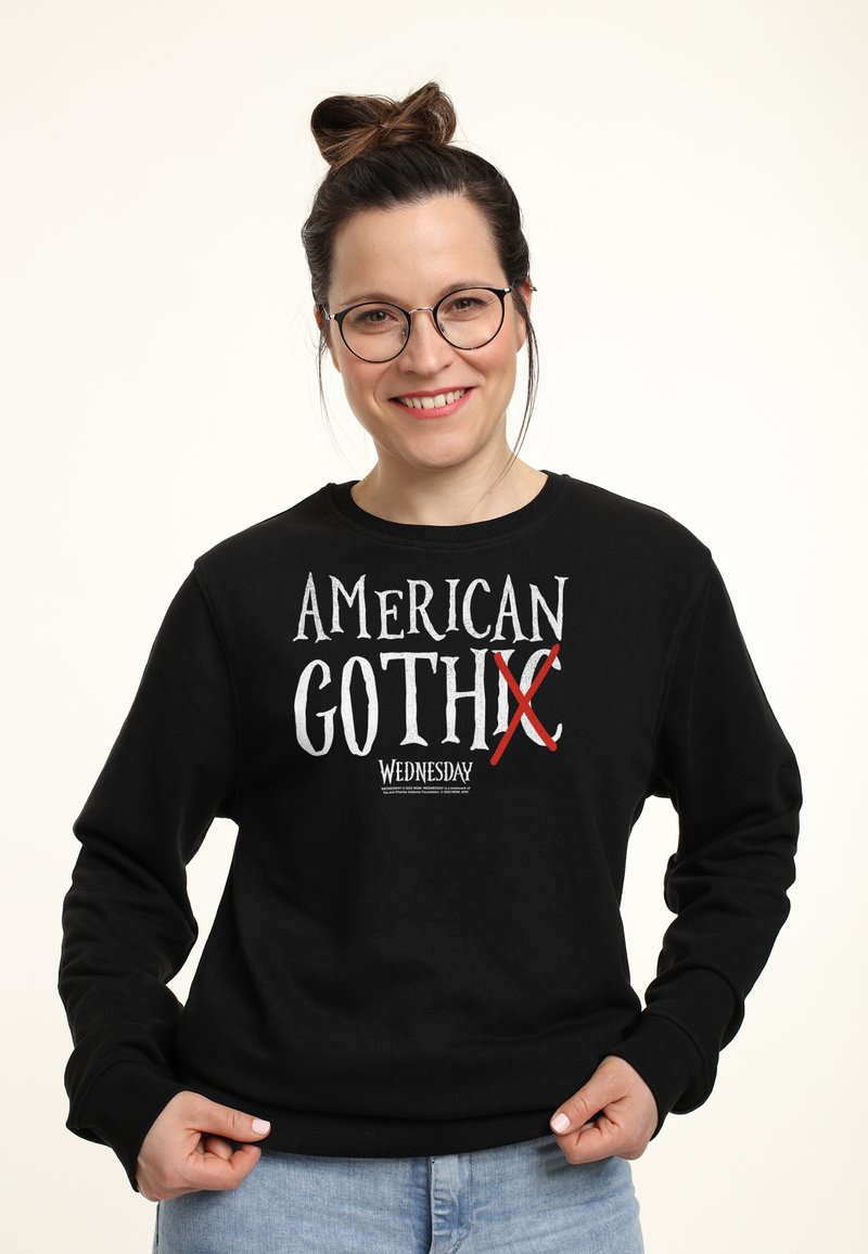 Donna sorridente con occhiali in un felpa nera con la scritta "American Goth X Wednesday," abbinata a jeans azzurri su uno sfondo bianco.