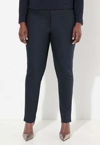 Marineblauwe slim-fit broek met een gladde textuur, subtiele frontnaad en elastische tailleband; gecombineerd met metallic puntige hakken.