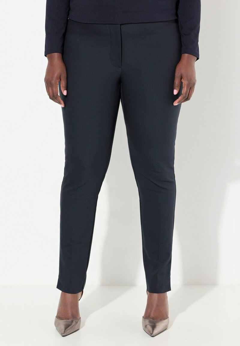 Marineblauwe slim-fit broek met een gladde textuur, subtiele frontnaad en elastische tailleband; gecombineerd met metallic puntige hakken.