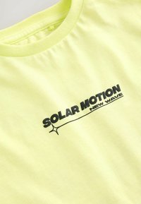 Kortærmet, iøjnefaldende neon gul T-shirt med rund hals og sort trykt tekst, der læser "SOLAR MOTION NEW WAVE." Blødt bomuldsstof.