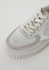 Zapatillas de ante gris claro con acentos trenzados y detalles plateados. Presenta una suela blanca gruesa y cordones planos y texturizados.