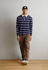 Polo de mangas longas azul-marinho com riscas brancas e colarinho branco, combinado com calças castanhas e sapatos azul-marinho, de pé num chão de madeira.