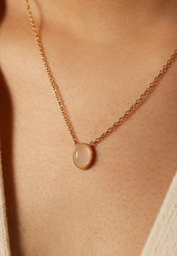 Gouden ketting met een ovale rozenkwarts hanger met een getextureerde bezel, close-up op de huid, met nadruk op detail en materiaal.