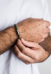 Žalias pynimas apyrankė su blizgiu sidabro metaliniu akcentu. Tekstūra yra lygi ir nupinta, pabrėžianti paprastą, minimalistinį dizainą.