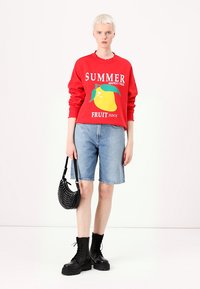 Sudadera roja con el texto "SUMMER" y un gráfico de mango amarillo, combinada con shorts de mezclilla azules, botas de combate negras y un bolso con tachuelas.