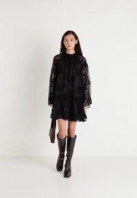 Crna prozirna haljina s cvjetnim vezom, dugim rukavima i volanima na rubu. Uparena s smeškim sjajnim čizmama do koljena i odgovarajućom torbom.