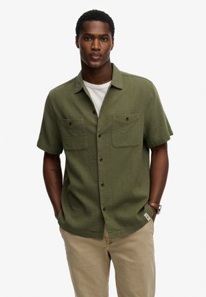 Uomo con camicia verde oliva a maniche corte abbottonata sopra una t-shirt bianca, pantaloni beige e orologio marrone, in piedi con le mani nelle tasche.