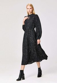 Robe noire midi avec un motif floral, manches bouffantes longues, col montant et détail en clef. Portée avec des bottines noires. Texture de tissu lisse.