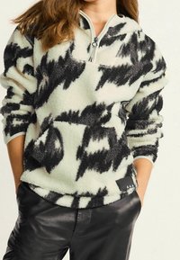 Fleece-pullover i creme med sorte abstrakte mønstre, halvlukning med lynlås og lange ærmer. Har en tekstureret blød stof og logo detaljer i siden.
