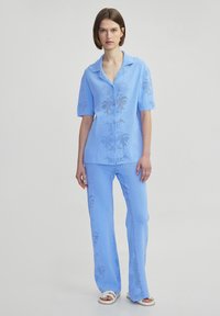 Světle modrý dvoudílný outfit se zkráceným rukávem a rozšířenými kalhotami, zdobený jemným květinovým vzorem a strukturovanou tkaninou.