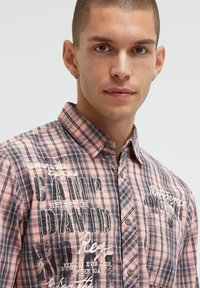 Karierte Kurzarmbluse in Pink und Grau. Mit Knöpfen, einem Kragen und grafischen Textmustern auf der Vorderseite. Aus einem leichten Stoff gefertigt.