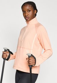 Ανοιχτόροζ πουλόβερ με φερμουάρ, με υφή σχεδίου, που διαθέτει ψηλό γιακά και διακριτικό λογότυπο. Χέρια κρατούν μπαστούνια πεζοπορίας με γκρι λαβές.