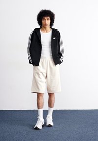 Corta-vento preto com fecho, listras brancas, t-shirt branca, calções beiges e ténis brancos. Modelo em pé contra um fundo simples.