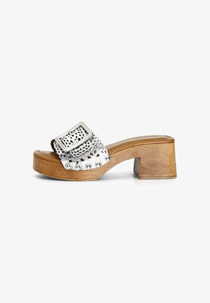 Stříbrná metalická slide sandál s širokým páskem s výřezy a ozdobnými hroty, spočívající na hnědém dřevěném bloku.