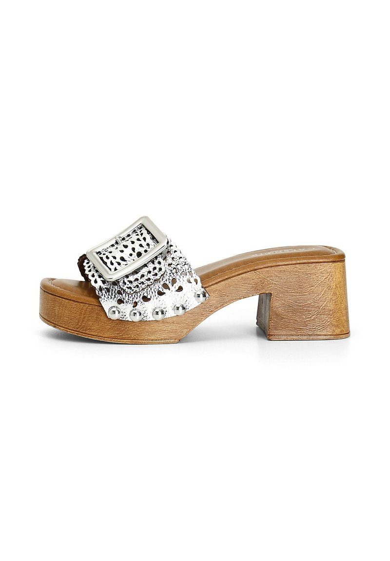 Sandal metalic argintie, cu o curea lată ce prezintă decupaje și studs decorative, așezată pe un toc din lemn bej.