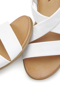 Weiße Sandalen mit zwei breiten Riemen, die sich über eine beige, strukturierte Fußbett kreuzen. Die Sohle ist flach und leicht flexibel, aus Gummi gefertigt.